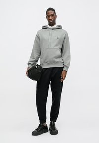 Siva dukserica s kapuljačom, prednjim džepom i pertlama, uparena s crnim trenirkama i crnim patikama, drži malu tamnu torbu.