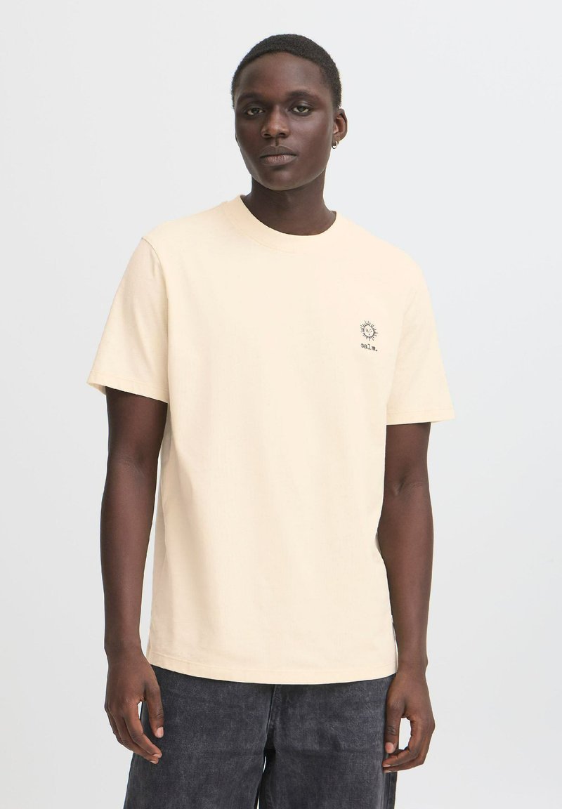 Lys beige bomulds t-shirt med korte ærmer, en rund halsudskæring og en solgrafik broderet på venstre bryst. Kombineret med mørke bukser.