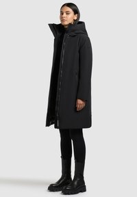 Naine kannab pikka musta sooja talvekatet kapuutsiga, kombineerituna mustade retuusidega ja massiivsete mustade saapaga.