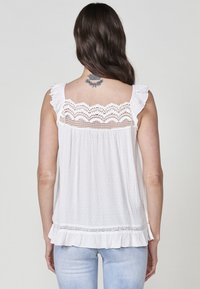 Haut sans manches blanc avec des bretelles à volants, détail en dentelle au dos et tissu texturé. Présente une forme évasée et un ourlet festonné.