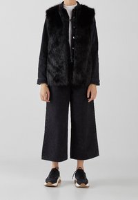 Person som bär en svart fuskpälsväst över en svart denimjacka, vida svarta avklippta byxor, vit skjorta och svarta sneakers med vita sulor.