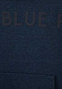 Temno modra teksturirana tkanina z velikimi črnimi črkami, ki pravijo "MODRA" in horizontalnim šivom blizu dna.