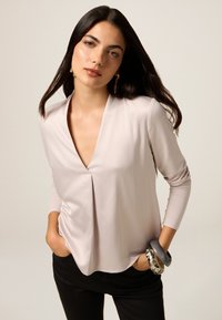Maglietta a maniche lunghe beige chiaro con scollatura a V profonda e parte anteriore plissettata, abbinata a pantaloni neri e bracciali chunk multi-colorati.