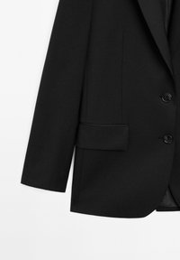 Blazer noir sur mesure avec deux boutons, une poche à rabat sur le côté droit et des manches longues sur fond blanc.