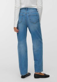 Donna che indossa jeans dritti azzurri chiaro, una maglietta bianca a maniche lunghe e ballerine nere, in piedi con la schiena rivolta verso la fotocamera.