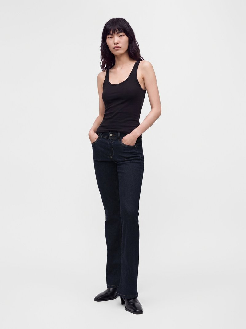 Femme aux longs cheveux noirs portant un débardeur noir, un jean bleu foncé et des chaussures noires, debout les mains dans les poches devant un fond uni.