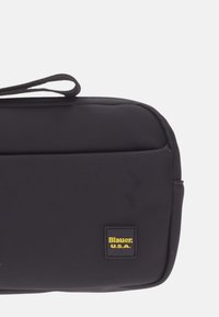 Fodero in nylon nero con una texture liscia, dotato di una maniglia superiore e una patch gialla con il logo. Forma rettangolare compatta. Nessun ulteriore motivo o dettaglio.