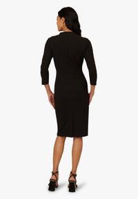 Robe noire, longueur genou, avec des manches trois-quarts, fermeture éclair au dos et fente ; présente une silhouette lisse et ajustée. Sandales noires à talons hauts.