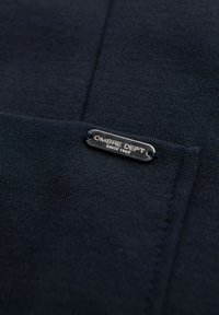 Mörk marinblått tyg med en liten metallskylt som läser "OMBRE DEPT SINCE 1989" sydd på en ficka.