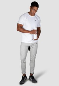 Hvid t-shirt med korte ærmer og et lille logo, grå sweatpants og sorte sportssko. Holder en hvid vandflaske. Enkel, funktionel design.
