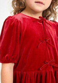 Een kind in een rode fluwelen jurk met pofmouwen en drie bijpassende fluwelen strikjes aan de voorkant.