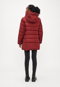 Rdeča puffer jakna s kapuco, prešita tekstura in nekoliko daljši hrbet. Oblečena z črnimi oprijetimi hlačami in debelimi črnimi čevlji.