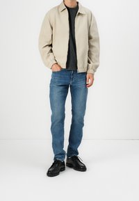 Chaqueta bomber beige claro con puños elásticos, usada sobre una camiseta oscura. Combinada con vaqueros azul medio y zapatos negros con cordones.