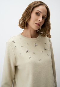 Beżowy sweter z okrągłym dekoltem, ozdobiony różnej wielkości przezroczystymi cyrkoniami rozmieszczonymi na przodzie, wykonany z miękkiego materiału.
