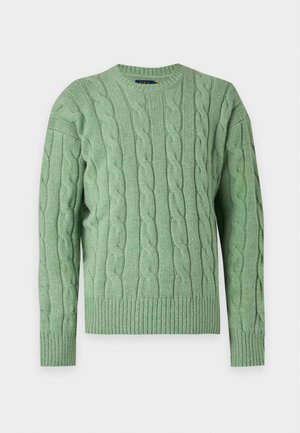 Ung mand iført en lysegrøn kabelstrik sweater over en mørkegrøn skjorte, lyseblå vide jeans og beige loafers, stående mod en hvid baggrund.