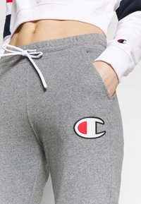Grå svettbyxor tillverkade av ett mjukt, strukturerat tyg. Har en vit dragkedja och en logotyp med röda och svarta detaljer.