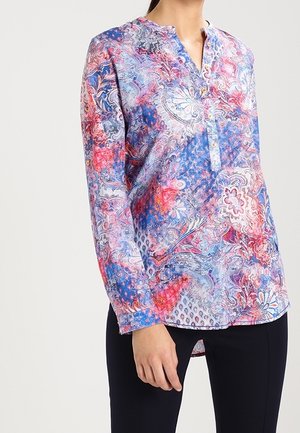 Bluzka z długim rękawem z zapinanym na guziki dekoltem w niebieskie, czerwone i białe wzory kwiatowe oraz paisley, noszona z czarnymi spodniami.