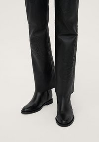 Bottes basses en cuir noir avec un bout arrondi et un petit talon carré, associées à un pantalon noir ajusté orné de motifs floraux embossés complexes.