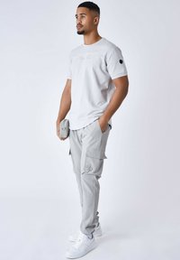 Camiseta de algodón gris claro con texto bordado, combinada con pantalones cargo gris claro que cuentan con bolsillos y un logo, complementada con zapatillas blancas.