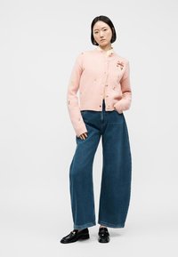 Γυναίκα που φοράει ροζ ζακέτα με κουμπιά και μικρά κεντημένα σχέδια, φαρδιά μπλε τζιν και μαύρα λοφάρια, στέκεται μπροστά σε μια απλή φόντο.