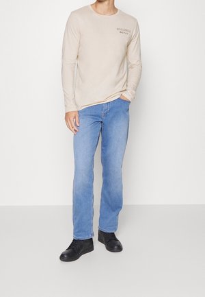 Beige Langarmshirt mit bedrucktem Design, kombiniert mit hellblauen Jeans. Schwarze Schuhe vervollständigen das legere Outfit.