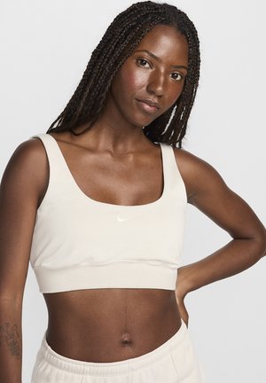Hellbeige Sport-BH mit quadratischem Ausschnitt, weichem Material und weißem Nike-Logo. Hoch geschnitten, zeigt den Bauch.