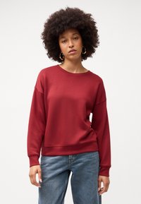 Felpa bordeaux con scollatura rotonda e spalle cadute, realizzata in tessuto morbido. Abbinata a jeans blu larghi, con polsini a costine delicati.