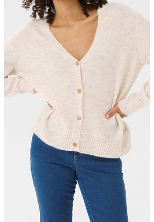 Femme portant un cardigan beige clair boutonné avec poignets et ourlet côtelés, associé à un jean bleu, mains posées sur les hanches.