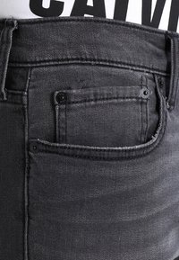 Grå denimjeans med fickdetalj, som har svarta knappar och en strukturerad tyg. Midjebandet har mörka sömmar och en slät finish.