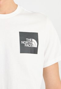 Balta medvilninė marškinėliai su juodu kvadrato logotipu ir baltu tekstu „THE NORTH FACE“, kurį puošia kreiva balta grafika kairėje krūtinės pusėje.