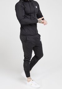 Svart idrotts-set med en figursydd zip-up hoodie och avsmalnande byxor. Har kontrasterande sidopaneler och broderad logotyp. Bärs med vita sneakers.