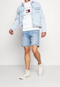 Chaqueta de mezclilla azul clara, sudadera blanca con capucha y logo, pantalones cortos de mezclilla azul clara y zapatillas beige con acentos rojos.