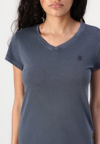T-shirt azul com decote em V, feito de algodão, com mangas curtas, um logótipo subtil no peito e um acabamento suave, ligeiramente texturado.