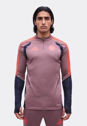 Jeune homme portant un haut de sport mauve avec des détails noirs et orange, debout avec une expression neutre sur un fond clair uni.