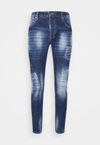 Slim-fit tummansiniset farkut, joissa on haalistuneita kohtia, kulutusta ja pieniä valkoisia sekä punaisia maalikastikkeita.