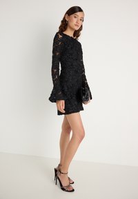 Melnas mežģīņu kleitas, kurai ir ziedu izšuvumi, garās piedurknes un izplešamas aproces. Apvienota ar mazu melnu rokassomu un siksnām augstpapēžu kurpēm.