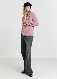 Maglione rosa chiaro e soffice con collo alto e maniche larghe, abbinato a jeans a gamba larga grigio scuro e scarpe nere, completato da un berretto nero.