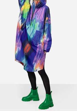 Persona con un impermeable con capucha multicolor con un patrón abstracto, leggings negros y botas tobilleras gruesas de color verde brillante.