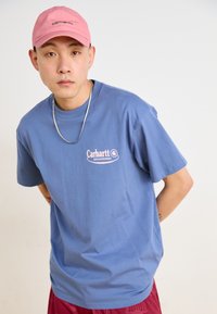 Šviesiai mėlynas medvilninis marškinėliai su rožiniu "Carhartt Delicatesse" logotipu, derinami su rožine kepure ir raudonomis šortais. Paprastas dizainas, apvalus kaklas.
