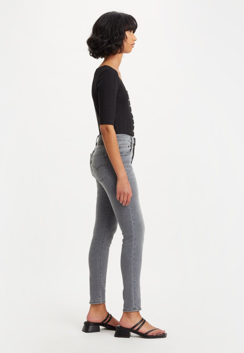 Graue, enge Jeans mit einem verblassten Finish, hoch tailliertes Design und klassischem Fünf-Taschen-Stil, kombiniert mit einem schwarzen gerippten Oberteil und schwarzen Sandalen.