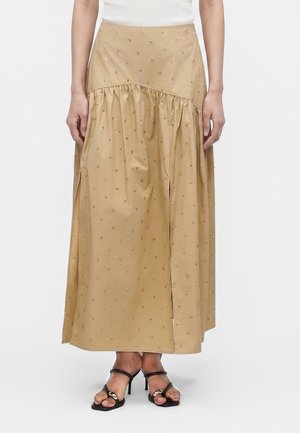 Beżowa spódnica midi z małymi zdobieniami w kropki, marszczonym szwem, rozcięciem z przodu, noszona z czarnymi sandałami na wysokim obcasie z paskami i białym topem.