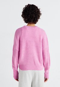 Chunkig rosa tröja med ribbat mönster och vida ärmar, med rundad hals och avslappnad passform, i kombination med gråa byxor.