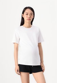 White cotton maternity t-shirt with short sleeves, slightly loose fit, round neckline, and side slits, paired with black shorts.