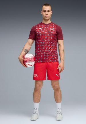 Tânăr în echipament sportiv roșu, ținând un balon de fotbal roșu și alb, stând în fața unui fundal gri simplu.