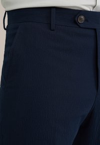 Pantaloni bleumarin dintr-un material texturat, cu buzunare laterale și închidere cu nasture, concepuți cu o croială elegantă și un tiv neted.