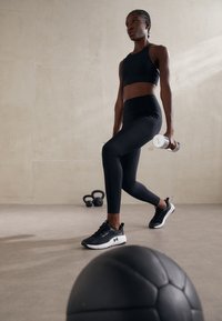 Juodas sportinis trumpas marškinėlis ir aukštai priglundančios leginsai, derinami su juodomis sportinėmis bateliais. Asmuo laiko dvi hantelius ir atlieka žingsnius į priekį. Juodas sporto kamuolys pirmame plane.