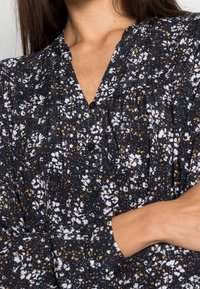 Czarna bluzka w kwiaty z białymi kwiatami i złotymi akcentami. Posiada dekolt w serek, detale z guzikami oraz bufiaste długie rękawy. Teksturowany materiał.