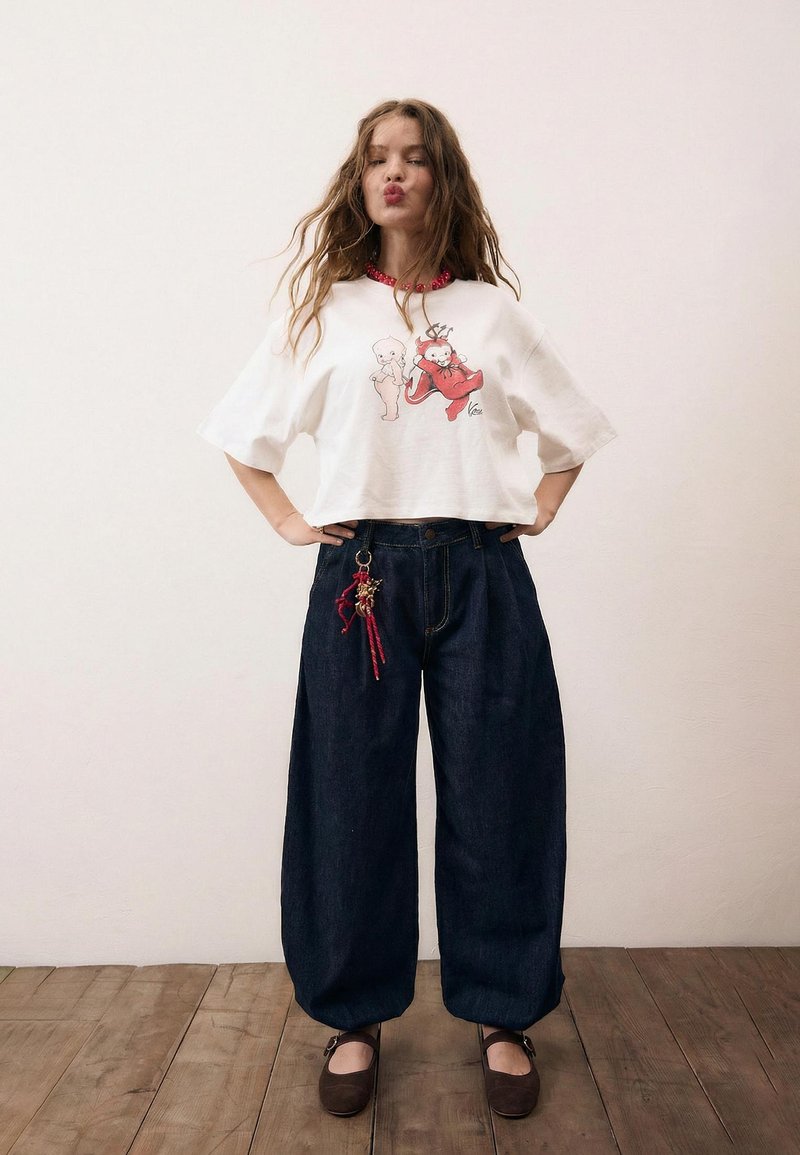 Frau mit langen, welligen Haaren, die ein weißes, grafisches Crop-Top, dunkle Jeans mit weitem Bein und einem roten Schlüsselanhänger sowie braune Mary-Jane-Schuhe trägt.