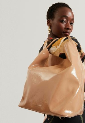 Donna con i capelli corti, che indossa orecchini d'oro e un top floreale, tiene una grande borsa beige lucida sulla spalla.