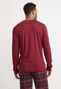 Camicia a maniche lunghe bordeaux con colletto rotondo, texture liscia e polsini aderenti, abbinata a pantaloni da pigiama a quadri nei colori rosso, nero e bianco.